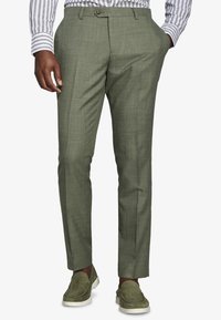 Hombre con pantalones de vestir a medida de color verde oliva y mocasines de ante a juego con suelas blancas, mano en el bolsillo, camisa de rayas blancas y grises.