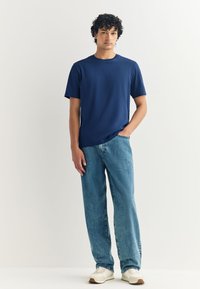 Tmavomodré tričko s krátkym rukávom a okrúhlym výstrihom, v kombinácii s svetlomodrými džínsami s rovnými nohavicami a bielymi teniskami s béžovými detailmi. Hladká textúra látky.