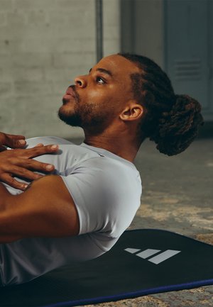 Athlete in a light grey fitted shirt performing an exercise on a black mat featuring white geometric logo, with a textured floor background.