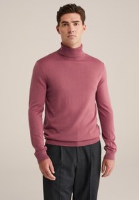 Lila pulover s turtleneck iz mehkega materiala, ki ima dolge rokave in fitted obliko, v kombinaciji s temno sivo krojenimi hlačami.