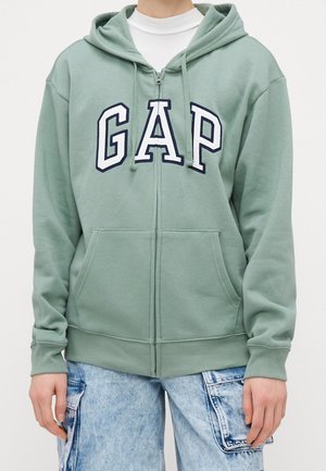 Personne portant un sweat à capuche zippé vert clair avec poche avant et lettres blanches "GAP,", associé à un jean cargo bleu clair.