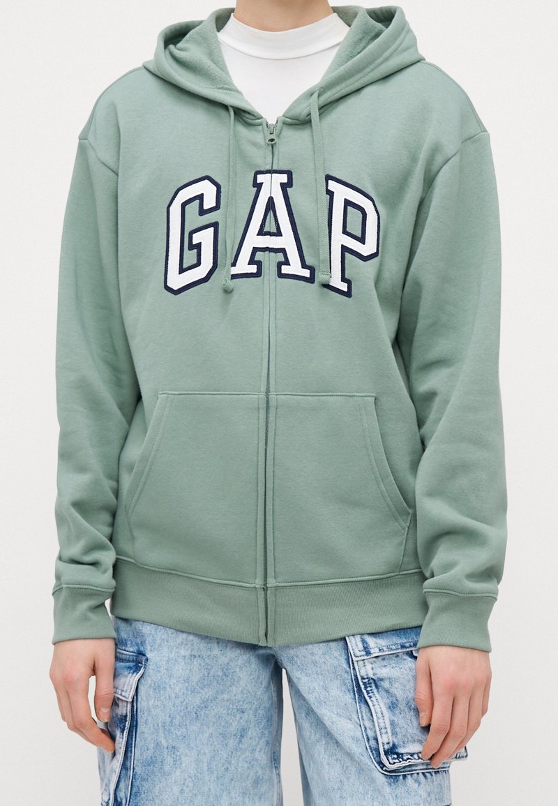 Personne portant un sweat à capuche zippé vert clair avec poche avant et lettres blanches "GAP,", associé à un jean cargo bleu clair.