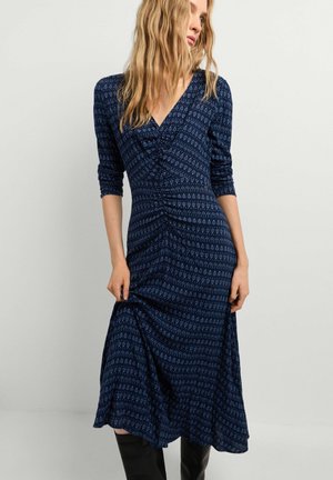 Mujer con cabello rubio ondulado que lleva un vestido midi azul marino de manga larga con estampado y botas negras, posando frente a un fondo liso y claro.
