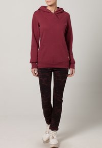 Bordowy hoodie z dekoltem w serek, kieszenią z przodu i beżowym guzikiem, w połączeniu z ciemnymi legginsami w wzory i białymi sneakersami.