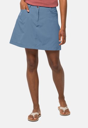 Vrouw met een blauwe knielange rok, een roze top en beige teenslippers, staand met één hand in haar zak tegen een effen achtergrond.
