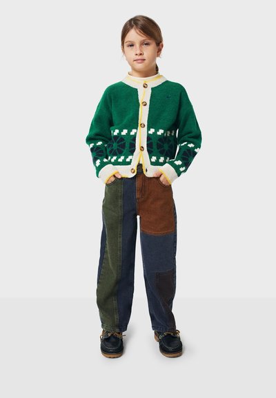 Vihreä neulecardigan, jossa on valkoisia yksityiskohtia ja nappikiinnitys, yhdistettyinä karkeakuvioisiin housuihin tummansinisessä, ruskeassa ja oliivinvihreässä. Mustat kengät täydentävät asua.