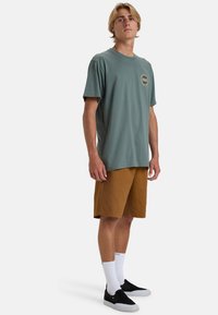 Hellgrünes T-Shirt mit kurzen Ärmeln und Grafikdruck auf der Brust, kombiniert mit braunen Shorts, weißen Socken und schwarzen Slippers.