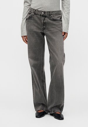 Personne portant un jean noir délavé à jambes larges, un haut gris à manches longues et des chaussures noires à bout carré, debout devant un fond uni.