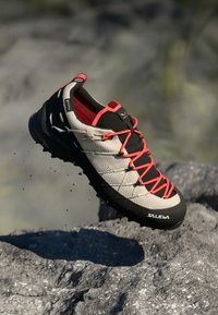 Leichtgewichtiger Wanderschuh mit einem oberen Teil aus beigem Textil, schwarzen Akzenten und roten Schnürsenkeln; robuste Sohle mit ausgeprägtem Profil für Trittfestigkeit.