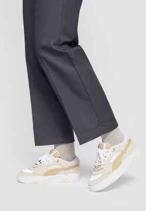 Person iført beige og hvide sneakers med lysegrå sokker og mørkegrå bukser med korte ben, stående på en lys baggrund.