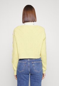 Maglione giallo a maglia con un motivo a rombi, design crop e polsini a coste. Indossato sopra jeans azzurri chiari, vista posteriore mostrata.