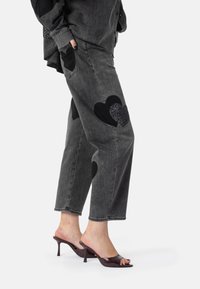 Čierne džínsy srdcového tvaru so záplatami z čierneho a trblietavého čierneho materiálu. Uvoľnený strih s piatimi vreckami a pretočeným lemom.