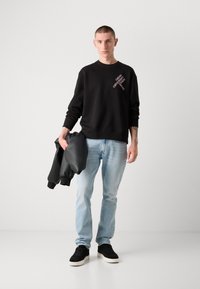 Hanorac negru cu text alb logo, blugi de culoare albastră deschisă, ținând o jachetă neagră. Poartă adidași negri cu talpă albă. Design simplu.