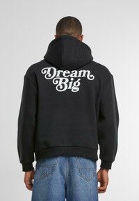 Fekete kapucnis pulóver, nagyméretű fehér "Dream Big" grafikával a hátsó oldalon, puha anyagból készült, állítható kapucnival és bordás mandzsettákkal.