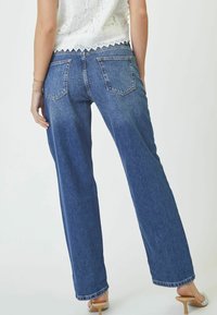 Breda denimjeans i blått med bakfickor och en lätt blekt yta, kombinerade med en vit spetsblus.