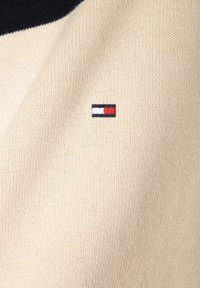 Strickpullover mit einer beigen Textur, einem marineblauen Kragen und einem kleinen gestickten Logo in Rot, Weiß und Blau an der Seite.