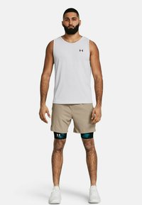 Weißes ärmelloses Sport-Shirt, beiges Shorts, schwarze und türkisfarbene Kompressionsärmel, kombiniert mit weißen Sportschuhen. Under Armour-Logo sichtbar.