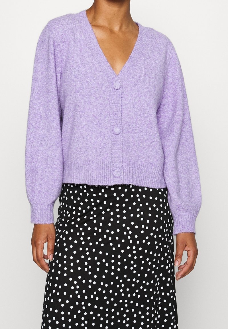 Lavendelfärgad kort tröja med V-ringning, knappförslutning och ribbade ärmmuddar. Matchas med en svart kjol med vita prickar.