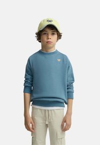 Niño con pelo rizado que lleva una gorra amarilla, una sudadera azul y pantalones cargo beige, de pie frente a un fondo liso.