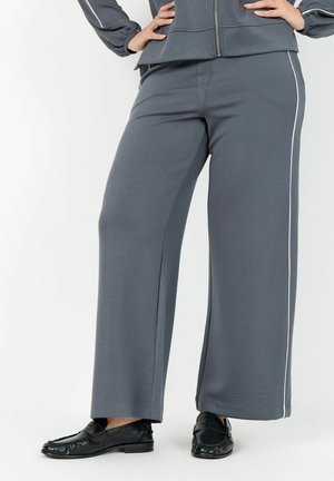 Person wearing wide-leg gray pants with white side stripe and black loafers, hands resting on hips against plain background.