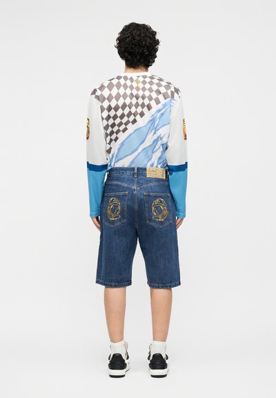 Άτομο με σγουρά μαλλιά φοράει μακρυμάνικο πουκάμισο με καρό και κυματιστά σχέδια, τζιν σορτς με μπαλώματα κράνους αστροναύτη στις πίσω τσέπες.