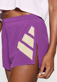 Μωβ αθλητικό σορτς με διάτρητη υφή, που διαθέτει μπεζ σχέδιο με τρεις ρίγες και πλευρικές σχισμές για καλύτερο αερισμό και κίνηση.