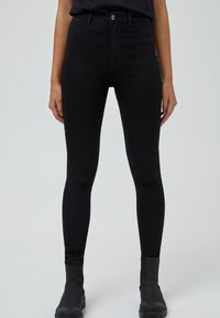 Jeans skinny negros de talle alto con una textura suave, diseño de cinco bolsillos y cierre de botón metálico, combinados con botas negras de tobillo.
