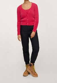 Chundelatý růžový kardigan s dlouhými rukávy a knoflíky, spárovaný s úzkými černými kalhotami a světlými šněrovacími botami s masivními podrážkami.