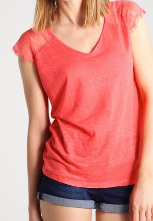 Kobieta ubrana w koralowy różowy top z dekoltem w serek, koronkowymi krótkimi rękawami i ciemnoniebieskie podwinięte krótkie spodenki jeansowe na jednolitym jasnym tle.