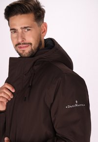 Brun hættejakke med lynlås, snore i hætten og et logo på ærmet. Glat, vandafvisende stof. Modellen smiler svagt.