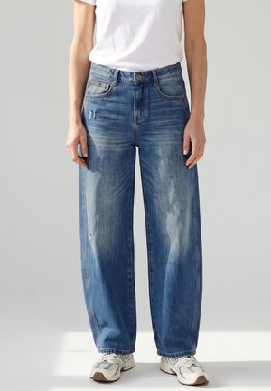 Persoon die een blauwe high-waist wijde broekspijp jeans draagt met lichte slijtage, gecombineerd met witte sneakers en een wit T-shirt met korte mouwen.