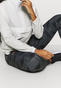 Veste gris clair à fermeture éclair avec un col montant, assortie à un legging noir à motif géométrique. Chaussures noires de sport.