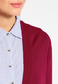 Red cardigan with a soft texture, open front, worn over a light blue and white striped collared shirt with button details.