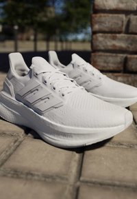 Coppia di scarpe da corsa sportive bianche con tessuto a maglia e tre strisce sul lato, posizionate su un pavimento in pietra all'aperto vicino a un muro di mattoni.