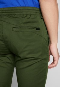Pantalon vert en tissu résistant, doté d'une ceinture élastique, d'une poche arrière et d'une étiquette logo noire sur le côté droit.