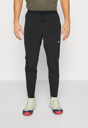 Homme portant un pantalon de running Nike noir avec une taille à cordon et des chaussures de running claires avec des semelles rouges et bleu marine.
