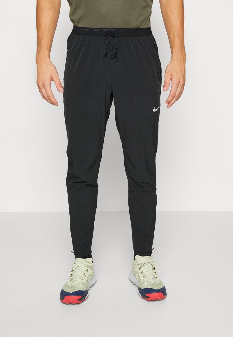 Homme portant un pantalon de running Nike noir avec une taille à cordon et des chaussures de running claires avec des semelles rouges et bleu marine.
