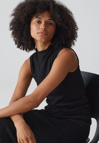 Zwarte mouwloze jurk met gestructureerde stof, hoge halslijn en aansluitende silhouet. Model zittend op een zwarte stoel, met natuurlijk krullend haar.