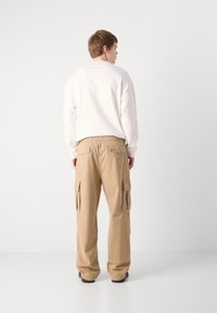 Beige cargo pants with elastic waistband, featuring side pockets and a loose fit. Worn with a plain white sweatshirt, viewed from the back.