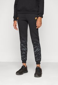 Sorte joggebukser med en oransje logo, med et gradient grå kamuflasjemønster langs nedre ben og ribbestrikkede ankelmansjetter.