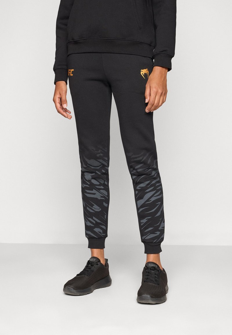 Sorte joggebukser med en oransje logo, med et gradient grå kamuflasjemønster langs nedre ben og ribbestrikkede ankelmansjetter.