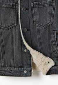 Chaqueta de mezclilla en gris oscuro con forro de piel sintética. Presenta dos bolsillos en el pecho, cierres de botón y costuras en contraste a lo largo de las costuras.