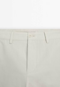 Gros plan sur la ceinture beige d’un pantalon avec un seul bouton fermé, des passants pour ceinture et une braguette zippée cachée.