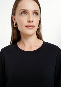 Crna dukserica s okruglim izrezom, glatke teksture i dugim rukavima. Model nosi zlatne naušnice u obliku obruča, prikazujući minimalistički stil.