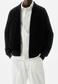 Giacca nera in corduroy con zip e colletto, abbinata a una camicia bianca con bottoni e pantaloni bianchi. Presenta tasche laterali e una vestibilità rilassata.