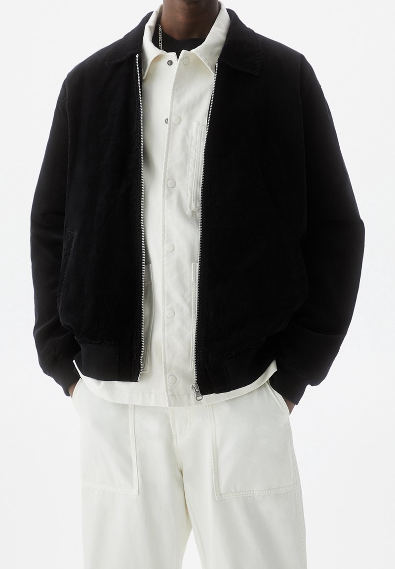 Schwarze Cordjacke mit Reißverschluss und Kragen, kombiniert mit einem weißen Hemd mit Knöpfen und weißen Hosen. Verfügt über Seitentaschen und einen entspannten Schnitt.