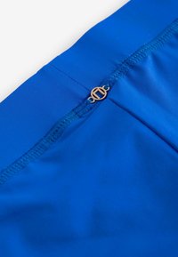 Blauwe zwemboxers van glad materiaal met een gouden embleemaccent en een veilige gestikte tailleband voor comfort en stijl.