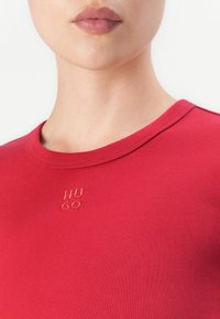 Tuvumā redzams cilvēks ar sarkanu kreklu ar apļveida kakla izgriezumu, uz kura ir diskrēti izšūts "HUGO" logo, kas atrodas centrā zem kakla izgriezuma.