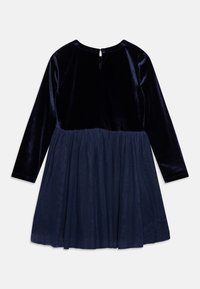 Marineblauwe jurk met een fluwelen bovenkant en een getextureerde tule rok. Lange mouwen en een opening met sleutelgat aan de achterkant. Zachte stof met een uitlopende pasvorm.