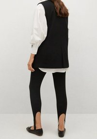 Schwarze ärmellose Blazer über einem weißen Shirt mit Puffärmeln, kombiniert mit schwarzen Leggings mit ausgeschnittenen Akzenten am Saum, getragen mit schwarzen Ballerinas.